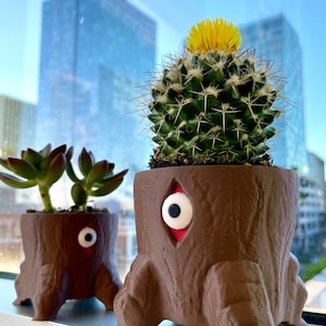 Pode incluir: Duas floreiras castanhas em forma de tronco de árvore com olhos arregalados, uma com uma suculenta e outra com um cacto com uma flor amarela. As floreiras estão numa janela com uma vista da cidade ao fundo.