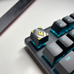 May include: Close-up of a custom keyboard keycap with a black base and a yellow and white design. The keycap is on a black keyboard with blue backlighting. Other black keys are visible.