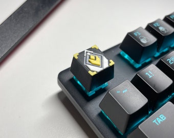 Könnte beinhalten: Nahaufnahme einer individuellen Tastaturtaste mit schwarzer Basis und gelb-weißem Design. Die Tastenkappe befindet sich auf einer schwarzen Tastatur mit blauer Hintergrundbeleuchtung. Weitere schwarze Tasten sind sichtbar.