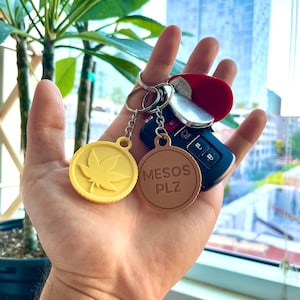 May include: A hand holding a set of keys and keychains. One keychain is yellow with a cannabis leaf design, and the other is brown with the text "MESOS PLZ". Also included are a car key and a red key fob.