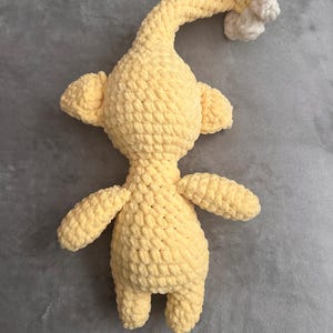 May include: A handmade, yellow crochet creature with a long, curved neck and small arms and legs. The head features a cluster of white crochet. The plush toy is made with a soft, textured yarn.