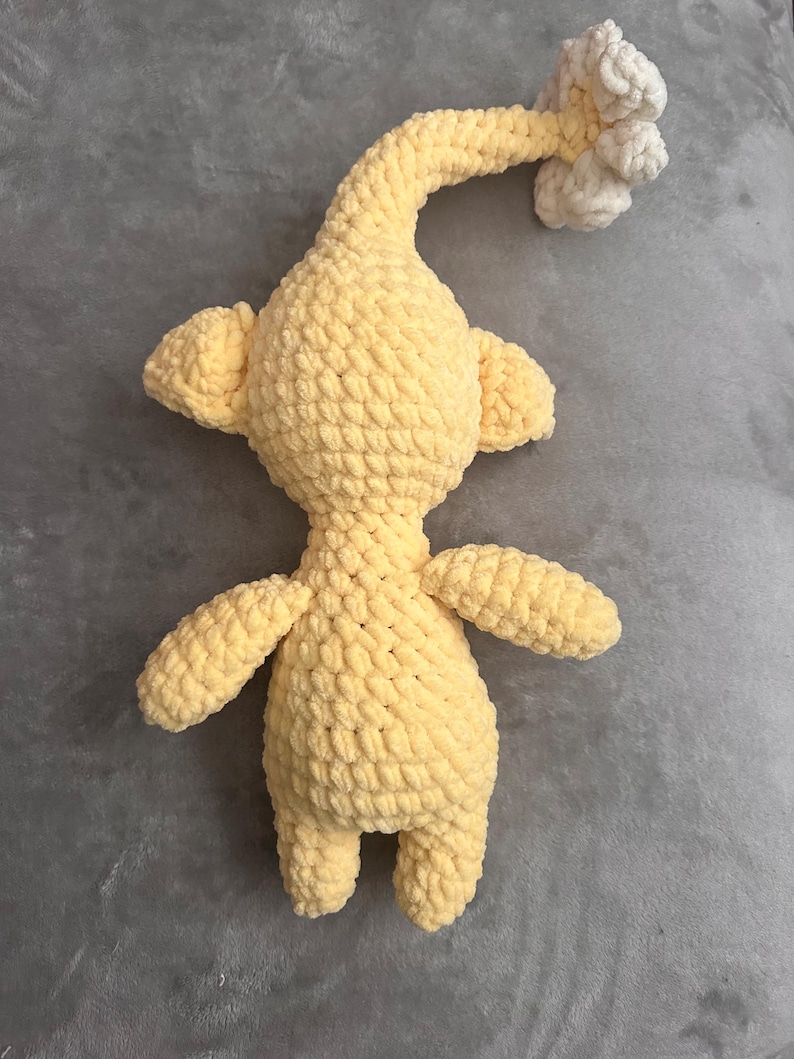 May include: A handmade, yellow crochet creature with a long, curved neck and small arms and legs. The head features a cluster of white crochet. The plush toy is made with a soft, textured yarn.