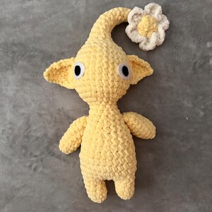 May include: A handmade, yellow crochet Pikmin plush toy with black eyes, small ears, and a curved stem. A white and yellow flower is attached to the stem. The plush is made of soft, textured yarn.