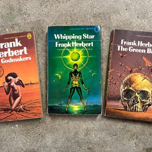 May include: Three vintage Frank Herbert paperbacks. The covers display illustrations: a figure in a desert, a futuristic figure, and a skull with insects. Titles are "The Godmakers", "Whipping Star", and "The Green Brain".