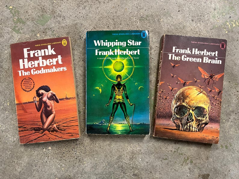May include: Three vintage Frank Herbert paperbacks. The covers display illustrations: a figure in a desert, a futuristic figure, and a skull with insects. Titles are "The Godmakers", "Whipping Star", and "The Green Brain".