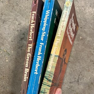 May include: Three vintage Frank Herbert paperbacks. The spines are visible, with titles including "The Green Brain," "Whipping Star," and "The Godmakers." The covers show signs of wear, with brown, blue, and orange colours.