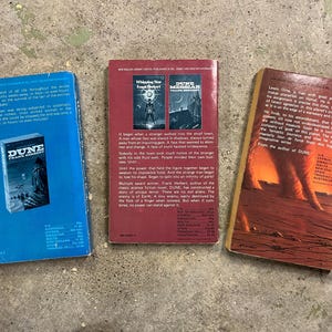 May include: Three paperback books are arranged on a concrete surface. The books are titled "Dune" by Frank Herbert, "Whispering Star" and "Dune Messiah" by Frank Herbert. The book covers display illustrations and text.