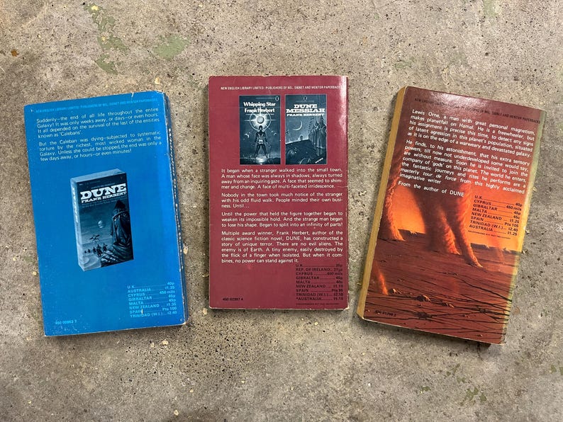 May include: Three paperback books are arranged on a concrete surface. The books are titled "Dune" by Frank Herbert, "Whispering Star" and "Dune Messiah" by Frank Herbert. The book covers display illustrations and text.