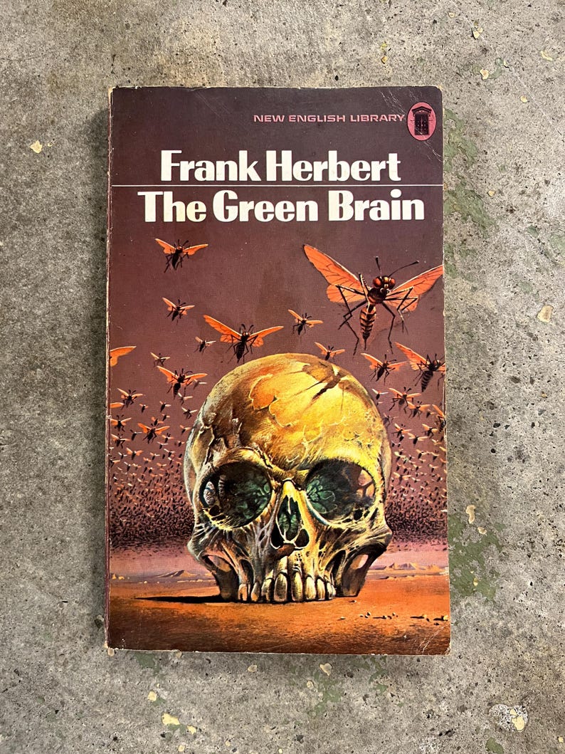 May include: A vintage paperback book titled "The Green Brain" by Frank Herbert. The cover shows a skull with green eyes and a swarm of insects with orange wings. The book is from the New English Library.