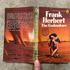 May include: A vintage paperback copy of "Frank Herbert The Godmakers" by Frank Herbert. The cover art depicts a desert scene with a kneeling figure. The book is open, displaying text and the continuation of the cover's artwork. The cover has a reddish-orange and brown color scheme.