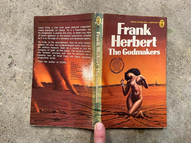 May include: A vintage paperback copy of "Frank Herbert The Godmakers" by Frank Herbert. The cover art depicts a desert scene with a kneeling figure. The book is open, displaying text and the continuation of the cover's artwork. The cover has a reddish-orange and brown color scheme.