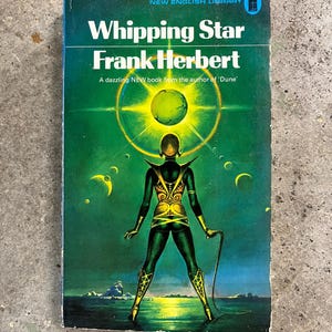 May include: Vintage paperback book cover for "Whipping Star" by Frank Herbert. The cover has a green backdrop with a figure in gold and green clothing holding a whip. The title and author's name are in white text.