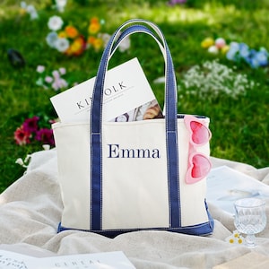 May include: A white canvas tote bag with navy blue trim and handles, embroidered with the name "Emma" in navy blue. The bag contains a magazine and pink heart-shaped sunglasses. The scene is set outdoors on a blanket with flowers.
