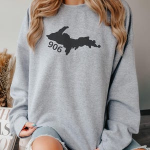 Puede incluir: Una sudadera gris con un contorno negro de la península superior de Michigan y el número 906 impreso en ella.