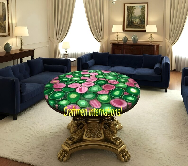 Puede incluir: Una mesa de centro redonda con una tapa de piedra de &aacute;gata verde y rosa. La mesa tiene una base dorada con detalles de cabeza de carnero. La habitaci&oacute;n cuenta con sof&aacute;s azules, cortinas color crema y una alfombra blanca.