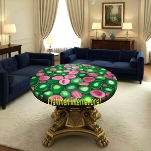 Puede incluir: Mesa de centro redonda con tapa de piedra de &aacute;gata verde y rosa. La mesa tiene una base dorada con detalles de cabeza de carnero. La habitaci&oacute;n tiene sof&aacute;s azules y cortinas blancas.