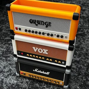 Op de afbeelding: Gestapelde miniatuurversterkerreplica's in oranje, wit en zwart. De bovenste versterker is oranje met de tekst "Orange", de middelste is wit met de tekst "VOX" en de onderste is zwart met de tekst "Marshall". Elk heeft een luidsprekerrooster en bedieningsknoppen.