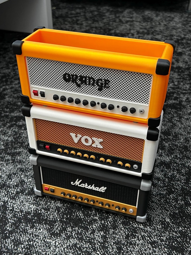Op de afbeelding: Gestapelde miniatuurversterkerreplica's in oranje, wit en zwart. De bovenste versterker is oranje met de tekst "Orange", de middelste is wit met de tekst "VOX" en de onderste is zwart met de tekst "Marshall". Elk heeft een luidsprekerrooster en bedieningsknoppen.