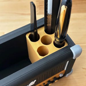 Op de afbeelding: Een zwarte en gouden bureau-organizer in de vorm van een Marshall-versterker. De organizer bevat een zwarte pen, een hobbymes en een zwart gereedschap. De voorkant van de organizer heeft het woord "Marshall" in het wit.