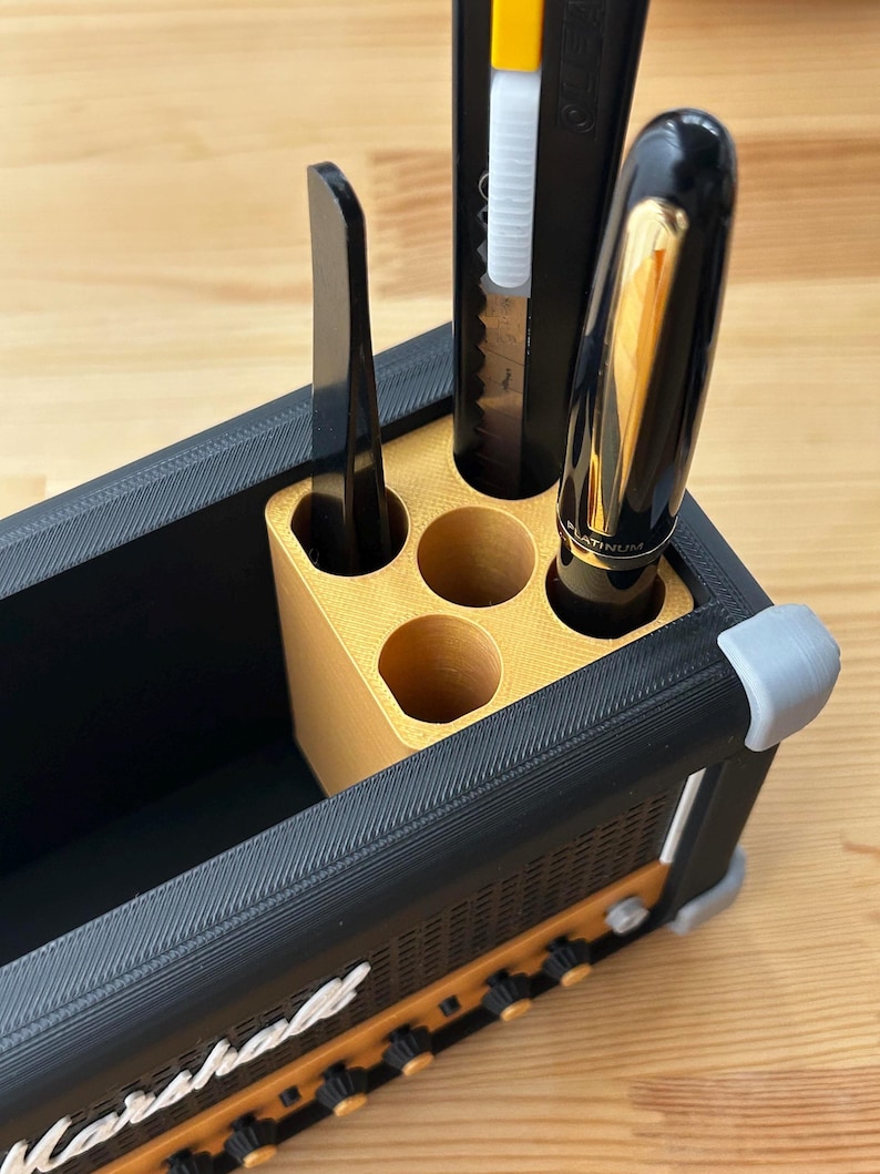 Op de afbeelding: Een zwarte en gouden bureau-organizer in de vorm van een Marshall-versterker. De organizer bevat een zwarte pen, een hobbymes en een zwart gereedschap. De voorkant van de organizer heeft het woord "Marshall" in het wit.