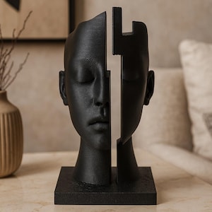 Könnte beinhalten: Eine schwarze Skulptur eines menschlichen Kopfes, vertikal geteilt, wobei eine Seite intakt und die andere eine geometrische Form ist. Die Skulptur steht auf einem quadratischen Sockel und ruht auf einer hellen Oberfläche. Der Kopf hat geschlossene Augen und eine glatte Textur.