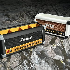 Op de afbeelding: Twee decoratieve dozen in de vorm van vintage gitaarversterkers. E&eacute;n is zwart met "Marshall" in wit, de andere is wit met "VOX" in bruin. Beide hebben knoppen en details van de luidsprekergrille.