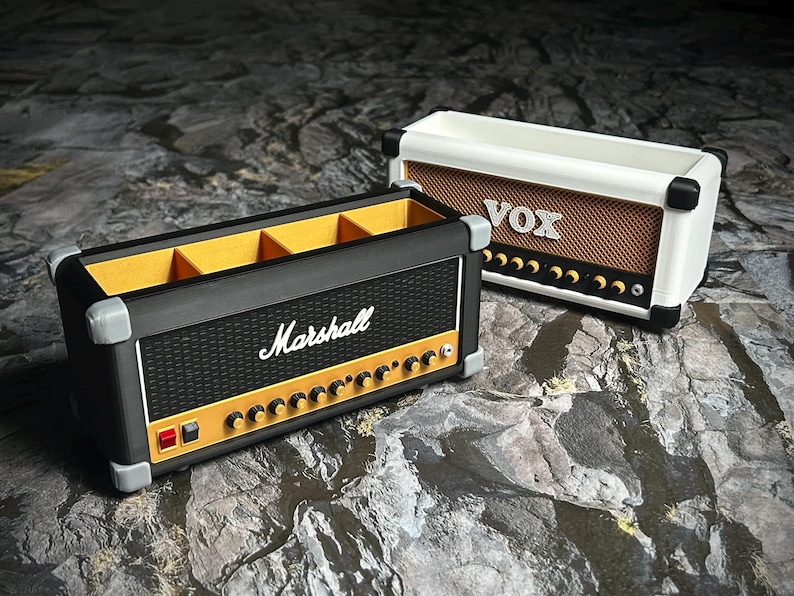 Op de afbeelding: Twee decoratieve dozen in de vorm van vintage gitaarversterkers. E&eacute;n is zwart met "Marshall" in wit, de andere is wit met "VOX" in bruin. Beide hebben knoppen en details van de luidsprekergrille.