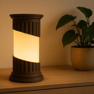 May include: A dark brown, column-shaped table lamp with a warm, glowing light. The lamp has a diagonal cut across the center. A potted plant with green leaves sits to the right of the lamp on a wooden surface.