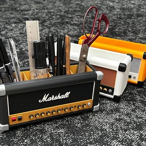 Op de afbeelding: Een zwarte bureau-organizer in de vorm van een Marshall-versterker, gevuld met pennen, potloden en een liniaal. De organizer heeft gouden knoppen en het Marshall-logo. Een wit-oranje versterker-vormige organizer staat ernaast, met een rood-handvat schaar.