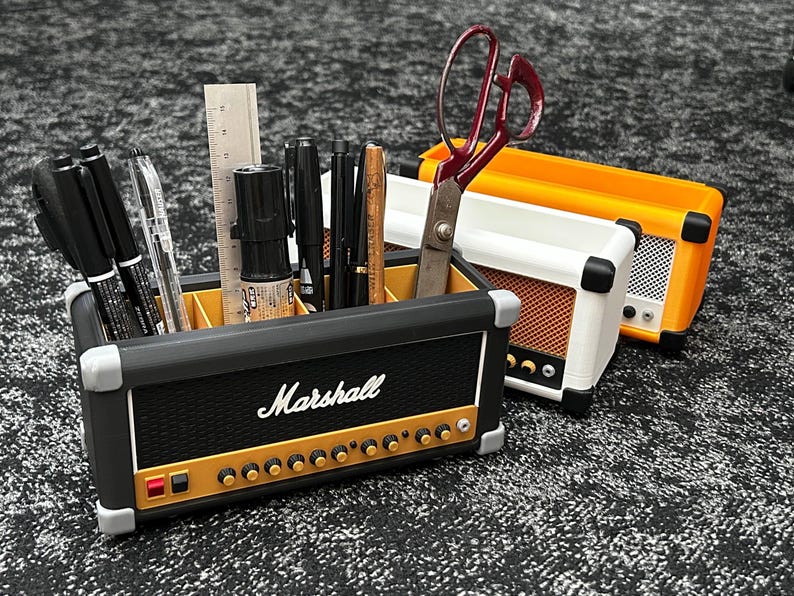Op de afbeelding: Een zwarte bureau-organizer in de vorm van een Marshall-versterker, gevuld met pennen, potloden en een liniaal. De organizer heeft gouden knoppen en het Marshall-logo. Een wit-oranje versterker-vormige organizer staat ernaast, met een rood-handvat schaar.