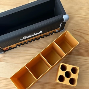 Op de afbeelding: Een zwarte en gouden decoratieve doos in de vorm van een Marshall-versterker, met het woord "Marshall" in witte letters. Er zijn ook drie gele opbergcontainers, een met vier compartimenten en een andere met zes gaten.