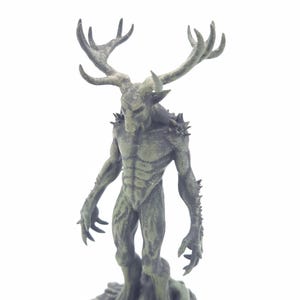 May include: A green figurine of a humanoid creature with deer antlers, sharp claws, and a muscular physique. The figure stands on a textured base, with a detailed, weathered appearance. The creature's face has a stern expression.