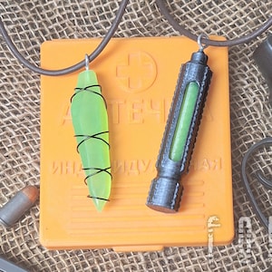 Può includere: Due collane con ciondoli in vetro verde. Un ciondolo è un flacone lungo, sottile, in vetro verde con un tappo nero. L'altro ciondolo è una forma a goccia di vetro verde con filo nero avvolto intorno. Le collane sono su un cordoncino in pelle marrone. Le collane sono sopra una scatola arancione con testo russo. Una pistola nera è sullo sfondo.