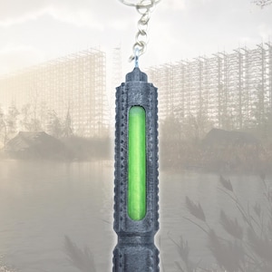 May include: A dark gray keychain with a cylindrical design and a bright green, glowing interior. The keychain is attached to a silver keyring. The background features a landscape with a body of water and a structure.