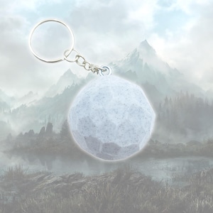 May include: A light gray, faceted sphere keychain with a silver-colored keyring and chain. The sphere has a speckled texture and is suspended against a blurred mountain landscape background.