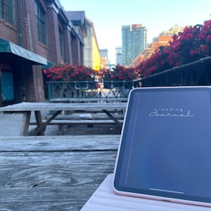 May include: A pink tablet with the words "reading journal" on the screen rests on a wooden picnic table. The tablet is in a pink case. The background includes a brick building, flowers, and a city street.