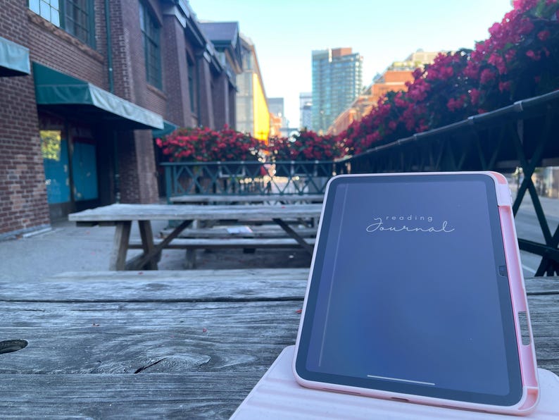 May include: A pink tablet with the words "reading journal" on the screen rests on a wooden picnic table. The tablet is in a pink case. The background includes a brick building, flowers, and a city street.