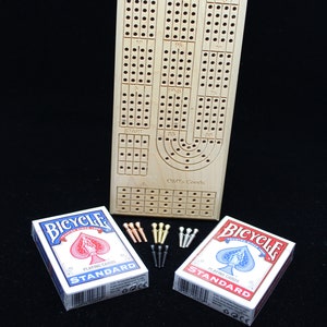 Op de afbeelding: Een houten cribbagebord met scoregaten, pinnen en de tekst "O&D's Goods". Twee kaartspellen van Bicycle en een set metalen pinnen zijn ook inbegrepen.