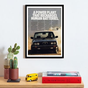 Könnte beinhalten: Gerahmte Vintage-Werbung mit einem schwarzen BMW-Auto, das auf einer unbefestigten Straße driftet. Die Anzeige enthält den Text "A POWER PLANT THAT RECHARGES HUMAN BATTERIES". Das Bild wird auf einer Holzoberfläche mit dekorativen Gegenständen angezeigt.