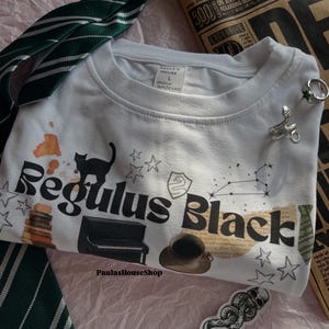 以下が含まれることがあります： 黒い猫、星、そして「Regulus Black」という文字が書かれた白いTシャツ。Tシャツには、小さなピアノ、コーヒーカップ、星座も描かれている。