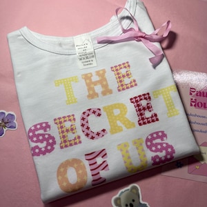 Puede incluir: Camiseta blanca con el texto "THE SECRET OF US" en letras de colores con patrones. Las letras están formadas por diferentes patrones, incluyendo estrellas, rayas y lunares.