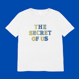 May include: White t-shirt featuring the text "THE SECRET OF US" in a decorative font. The text is in shades of yellow and blue. The shirt is made of a soft material and has a classic crew neck.