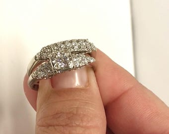 Könnte beinhalten: Ein silberner Ring mit einem großen, quadratischen Diamanten, der von kleineren Diamanten umgeben ist. Der Ring wird an einem Finger getragen.