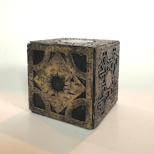May include: A decorative cube featuring a dark, weathered gold finish with intricate, etched geometric patterns and symbols on each face. The design includes a central circular motif surrounded by angular, engraved lines and dark recessed areas.