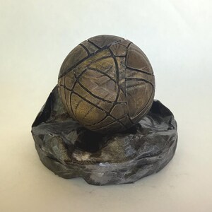 May include: A spherical object with a textured, segmented surface in shades of brown and gold, featuring dark recessed lines between the segments. The sphere rests within a dark, irregularly shaped, crinkled base with a metallic finish.