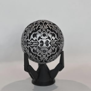 May include: A spherical object featuring a complex, raised silver filigree pattern over a black base, resting on a black tripod stand. The sphere has a horizontal seam running through the center, and the intricate design covers the entire surface.