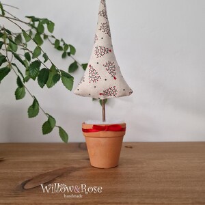 May include: A small, white fabric Christmas tree with a red and black print of Christmas trees. The tree is in a terracotta pot with a red ribbon around the top. The pot is sitting on a wooden surface. "Willow & Rose handmade" is written on the surface.