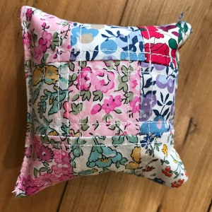 May include: A small, square, patchwork cushion with a floral print in shades of pink, blue, green, and red. The cushion is quilted with white thread and has a slightly ruffled edge.