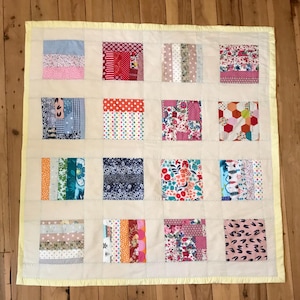 Puede incluir: Una colcha de retazos con fondo crema y borde amarillo. La colcha está compuesta por 16 cuadrados, cada uno con un patrón de tela diferente. Los patrones incluyen rayas, lunares, estampados florales y diseños geométricos. La colcha es una mezcla vibrante de colores, incluyendo rosa, rojo, azul, verde y amarillo.