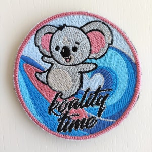 May include: A pink and blue embroidered patch with a cartoon koala riding a surfboard. The koala is gray with pink ears and a big smile. The text "Koala Time" is embroidered in black on the patch.